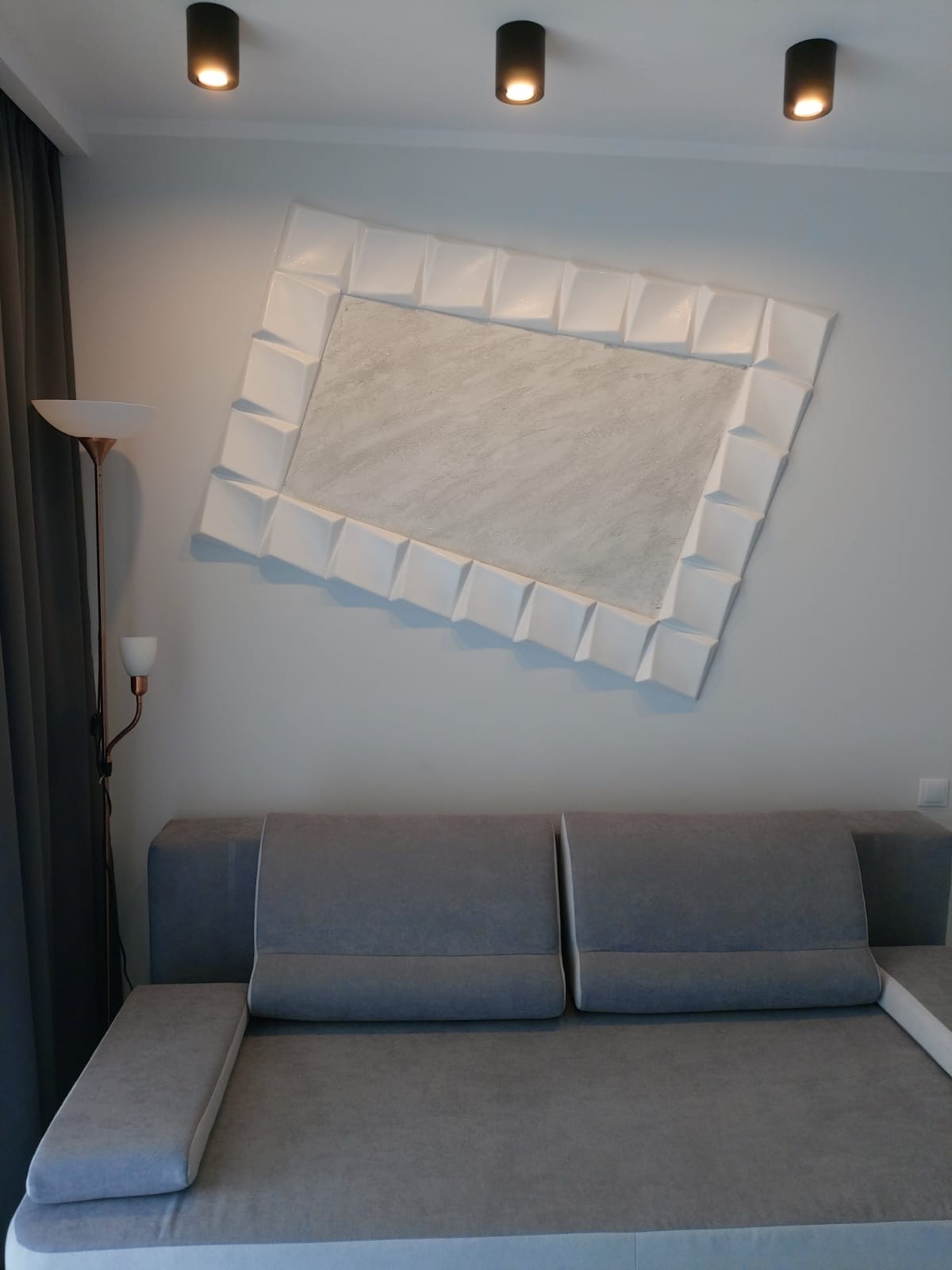 Nowoczesny salon z szarą sofą i dekoracyjną, białą ramą na ścianie, oświetloną trzema punktowymi lampami sufitowymi. Minimalistyczny design wnętrza.