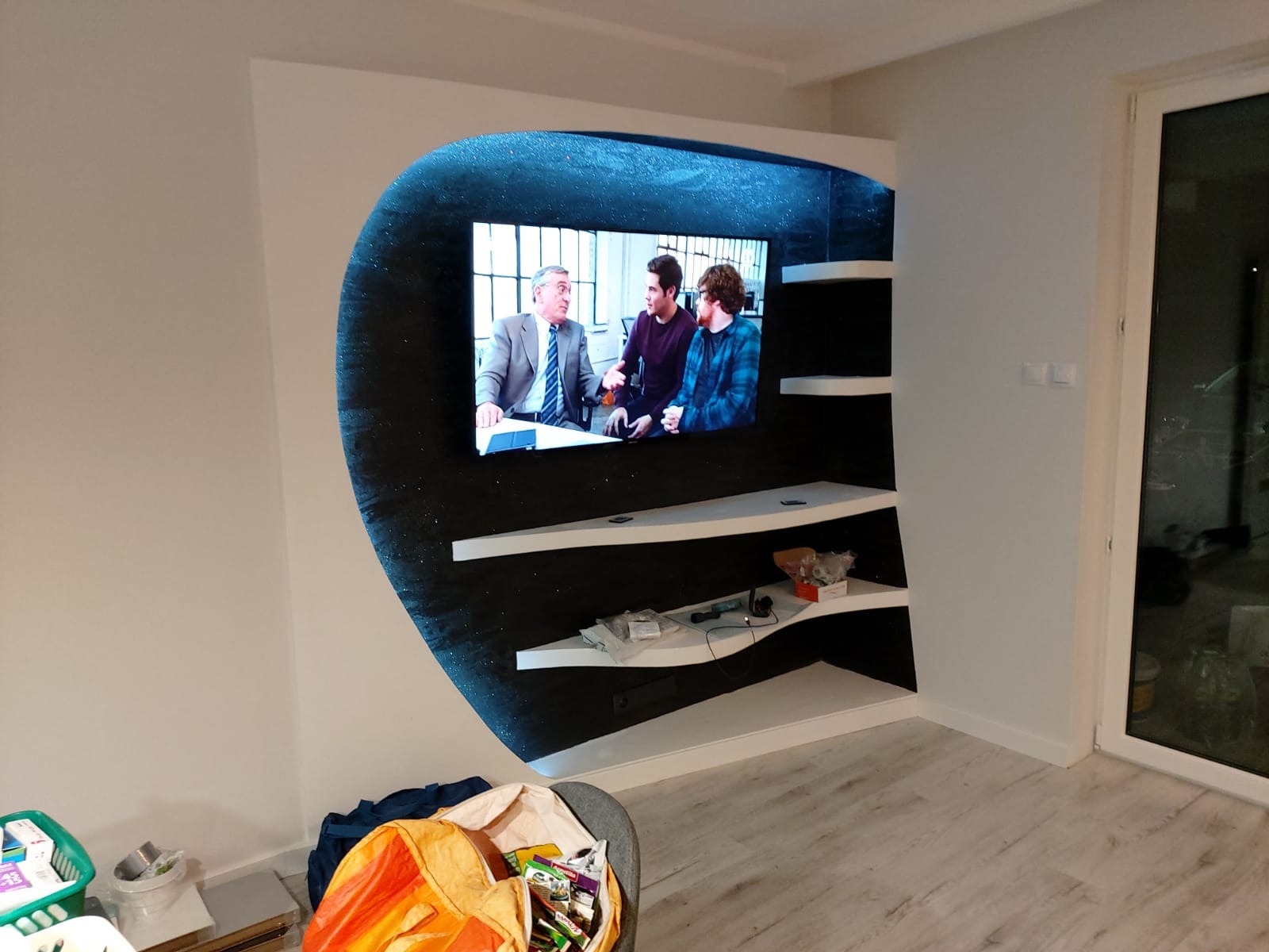 Nietypowa zabudowa ściany z podświetlaną wnęką na telewizor i półkami, wykończona w kolorach bieli i czerni z efektem brokatu. Widoczne elementy wykończenia wnętrza.