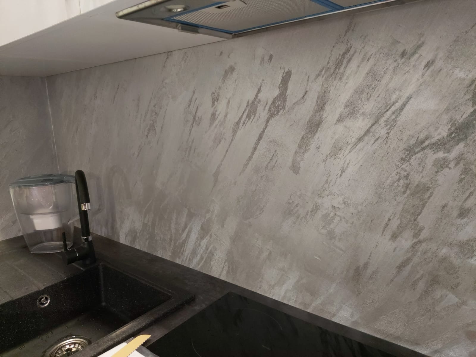 Ściana w kuchni wykończona dekoracyjnym tynkiem w odcieniach szarości, imitującym marmur. Widoczny fragment zlewu i płyty indukcyjnej. Nowoczesny design.