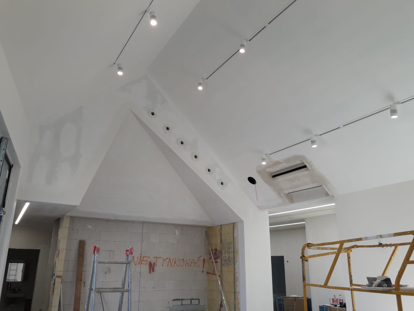 Biały sufit pod skosem z oświetleniem szynowym i punktowym. Widoczne otwory wentylacyjne i fragment ściany z napisem. Prace wykończeniowe w trakcie.