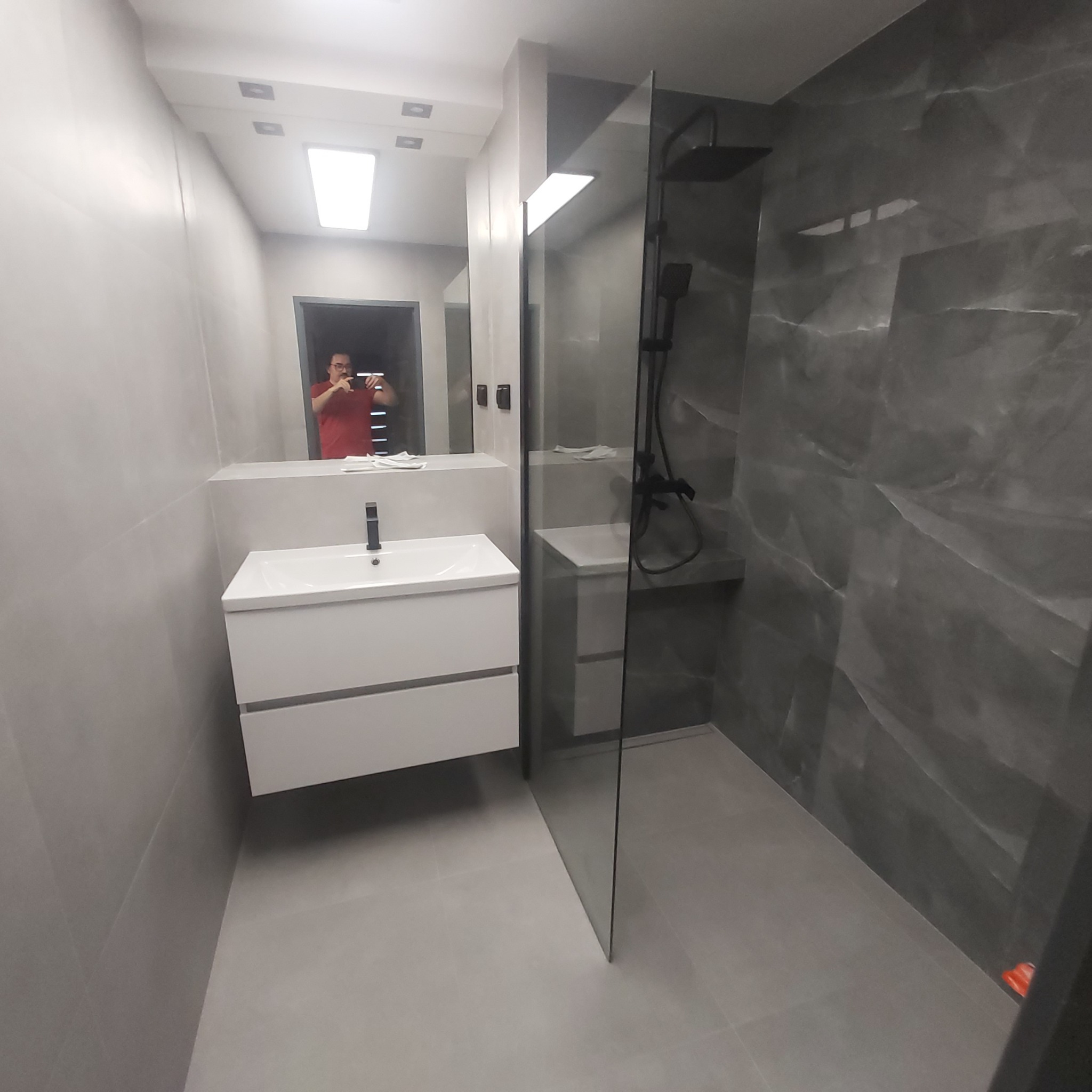 Nowoczesna łazienka po remoncie: szare płytki imitujące marmur, kabina walk-in z czarną armaturą i biała szafka z umywalką. W lustrze odbija się osoba robiąca zdjęcie.