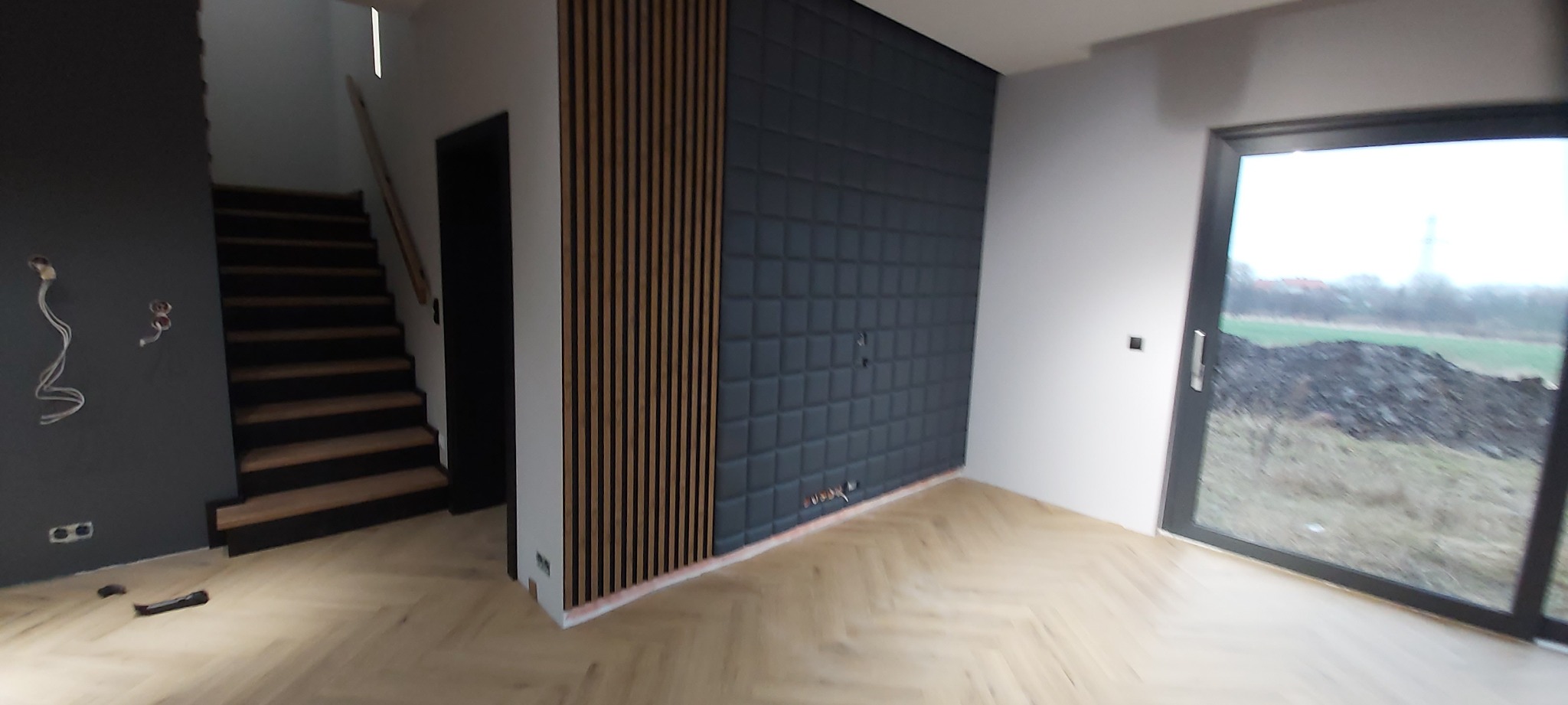 Nowoczesne wykończenie wnętrza w Głogowie: panel akustyczny w kolorze grafitowym, drewniane lamele i parkiet w jodełkę. Widoczne schody i duże okno.