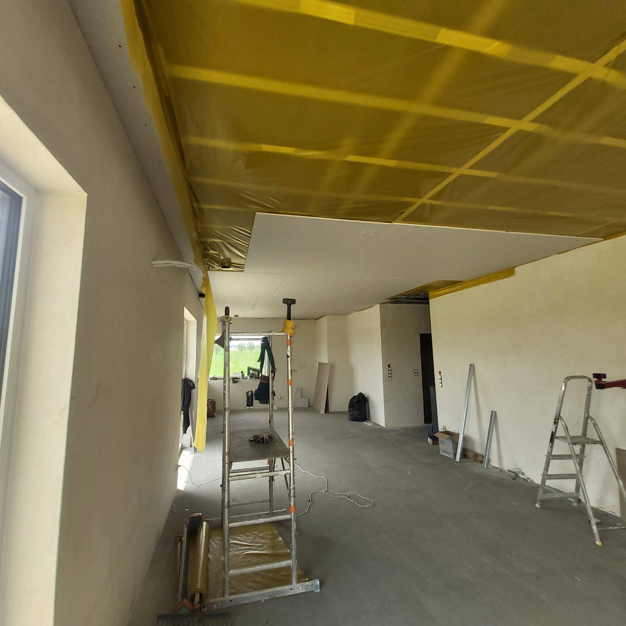 Montaż sufitu podwieszanego z płyt gipsowo-kartonowych w jasnym wnętrzu. Widoczny ruszt pod sufitem zabezpieczony folią i drabina monterska.