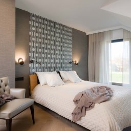 Firma JFK - Nowoczesna sypialnia na poddaszu z łóżkiem, wzorzystą tapetą i skórzanym fotelem z narzutą. Uwagę przykuwają lampki ścienne i parkiet ułożony w jodełkę.