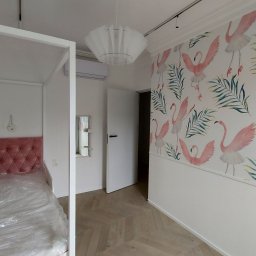 Firma JFK - Pokój po remoncie: łóżko z różowym zagłówkiem, ściana z motywem flamingów, parkiet w jodełkę. Widoczny klimatyzator i nowoczesna lampa.