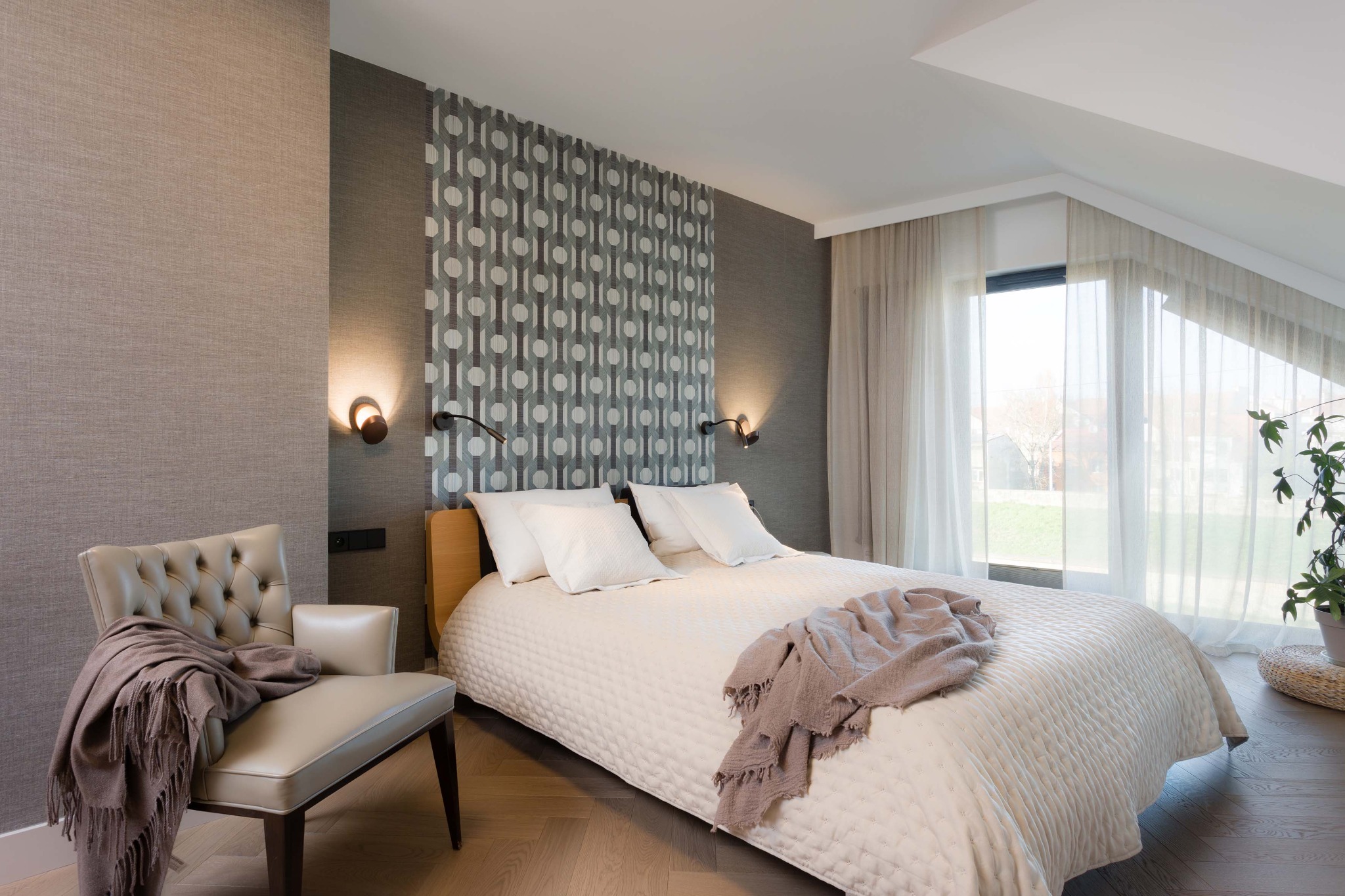 Nowoczesna sypialnia na poddaszu z łóżkiem, wzorzystą tapetą i skórzanym fotelem z narzutą. Uwagę przykuwają lampki ścienne i parkiet ułożony w jodełkę.