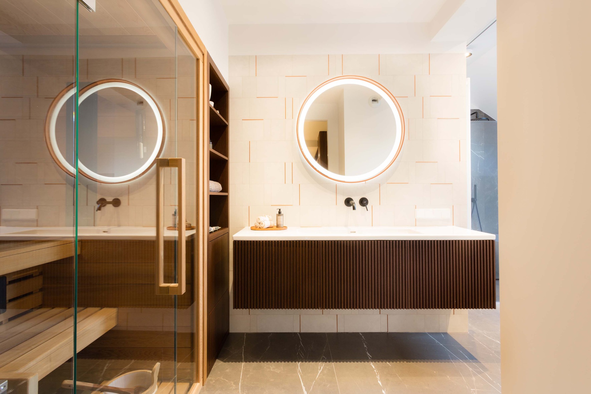 Nowoczesna łazienka z sauną: drewniana zabudowa umywalki z okrągłym lustrem LED, beżowe płytki i marmurowa podłoga. Widoczna sauna za szklaną ścianą.