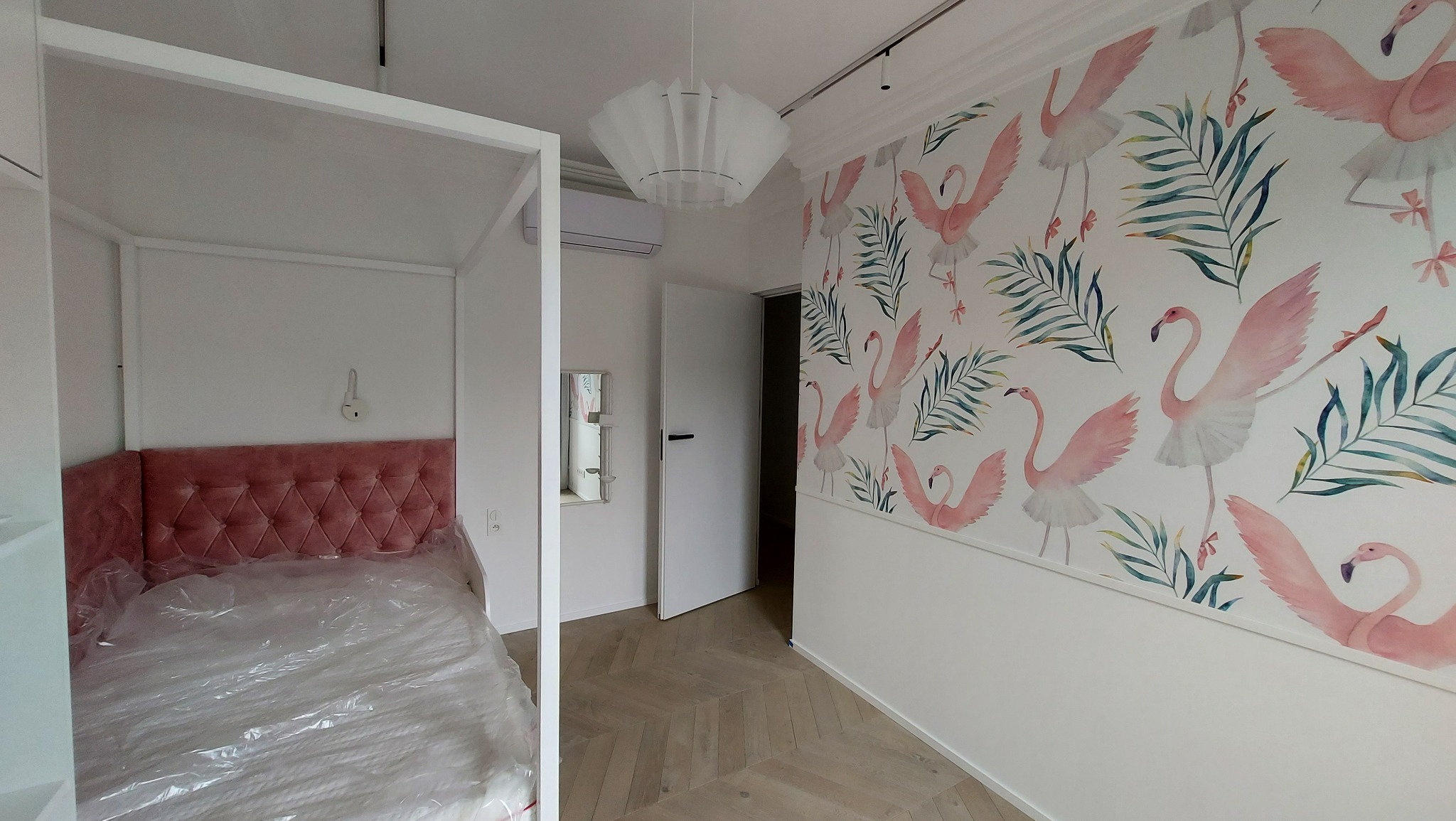 Pokój po remoncie: łóżko z różowym zagłówkiem, ściana z motywem flamingów, parkiet w jodełkę. Widoczny klimatyzator i nowoczesna lampa.