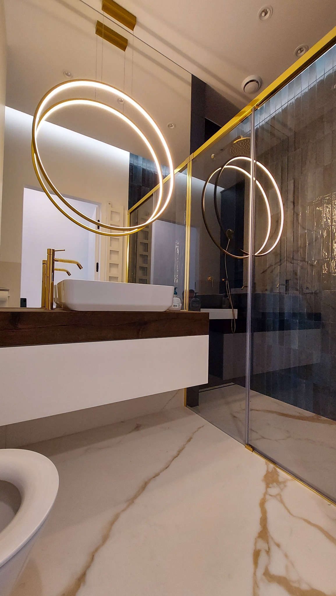 Nowoczesna łazienka z marmurową podłogą, złotą armaturą i oświetleniem LED w kształcie okręgów. Kabina prysznicowa ze złotymi profilami i ciemnymi płytkami.