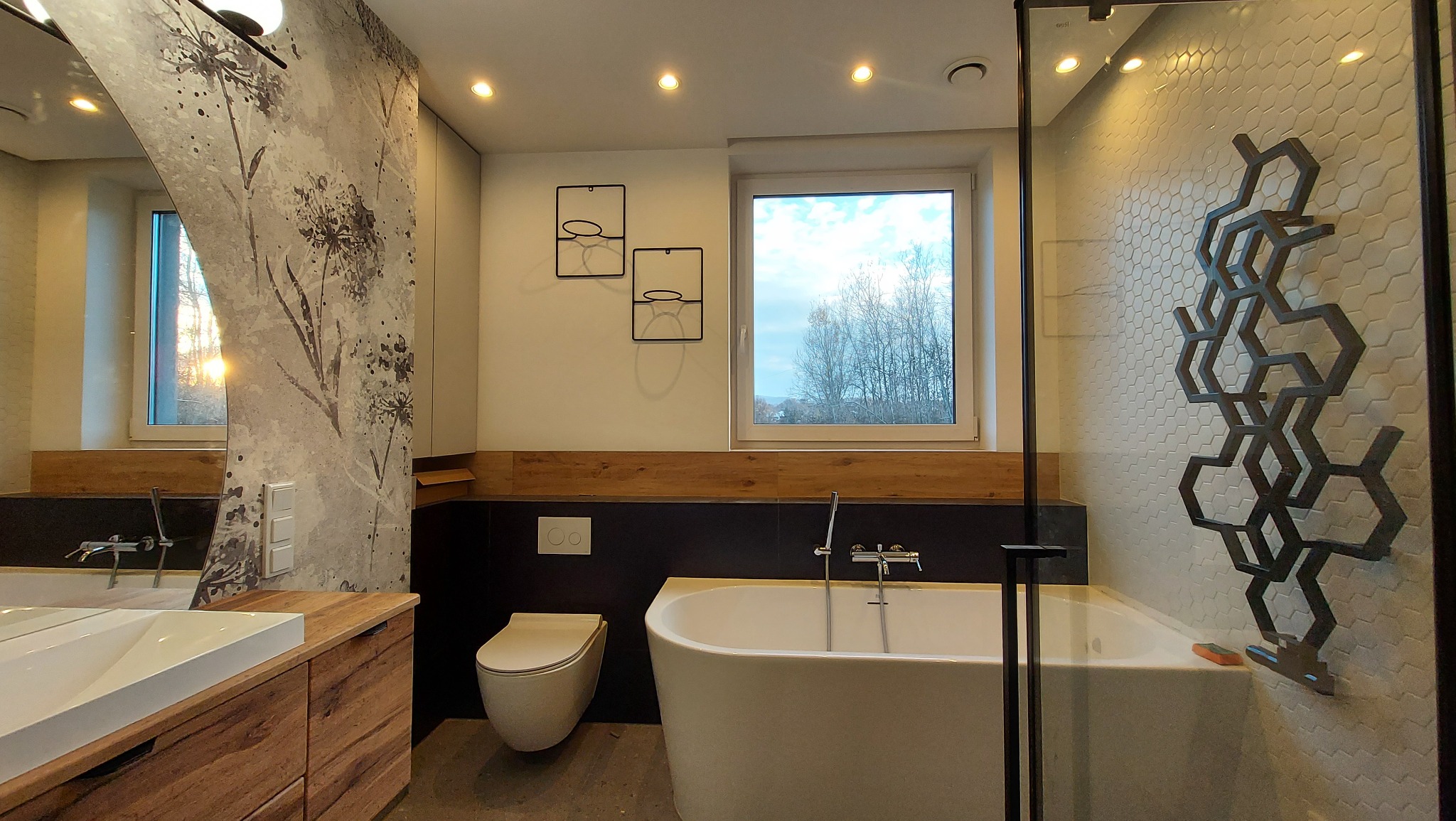 Nowoczesna łazienka z wolnostojącą wanną, umywalką na drewnianej szafce i dekoracyjnym grzejnikiem na heksagonalnej ścianie. Okno z widokiem na drzewa.