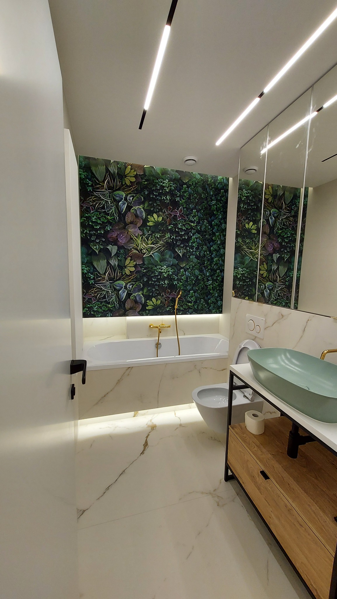 Nowoczesna łazienka z wanną, umywalką w odcieniach zieleni i ścianą z motywem roślinnym. Białe płytki z marmurowym wzorem na podłodze i ścianach.