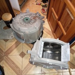 FIXIT SERWIS - Rozłożona obudowa pralki automatycznej na podłodze z narzędziami w tle, ukazująca etap demontażu podczas naprawy. Widoczne elementy bębna i obudowy.