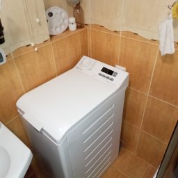 FIXIT SERWIS - Biała pralka Electrolux z górnym załadunkiem w łazience z brązowymi płytkami. Widoczny panel sterowania i fragment umywalki. Ujęcie z bliska, lekki kąt.