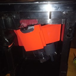 FIXIT SERWIS - Wnętrze ekspresu do kawy z widocznymi elementami konstrukcyjnymi i pomarańczowymi pojemnikami. Widoczne ślady użytkowania i demontażu.