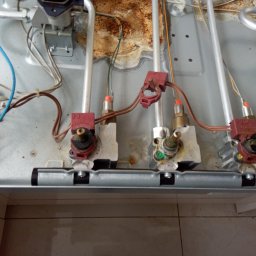 FIXIT SERWIS - Wnętrze kuchenki gazowej z widocznymi elementami instalacji gazowej i elektrycznej, w trakcie naprawy lub serwisu. Widoczne ślady użytkowania i zabrudzenia.