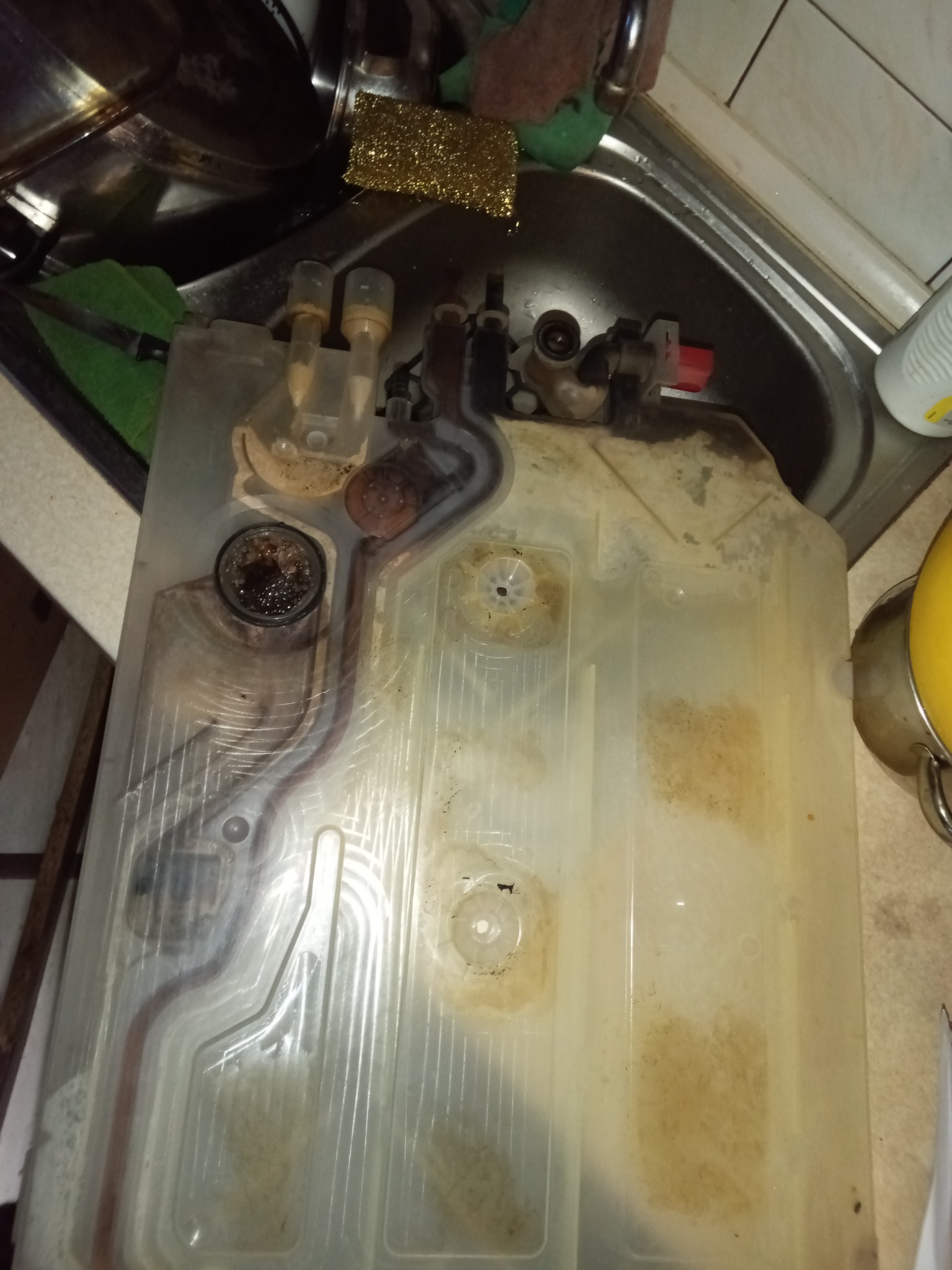 Wyjęty, zabrudzony element zmywarki w zlewie, widoczne osady i kamień. Elementy instalacji z tworzywa sztucznego i metalu. Widok z góry, ostre światło.