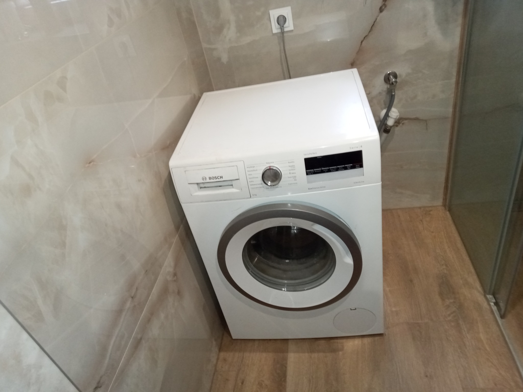 Biała pralka Bosch Serie 4, ustawiona w rogu łazienki z beżowymi płytkami imitującymi marmur i drewnianą podłogą. Widoczny wąż doprowadzający wodę i gniazdko elektryczne.