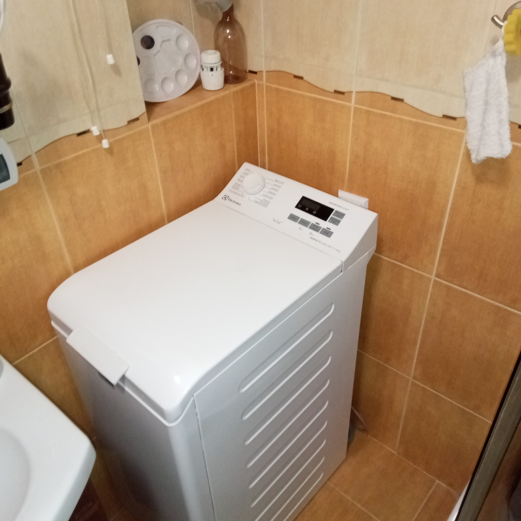 Biała pralka Electrolux z górnym załadunkiem w łazience z brązowymi płytkami. Widoczny panel sterowania i fragment umywalki. Ujęcie z bliska, lekki kąt.