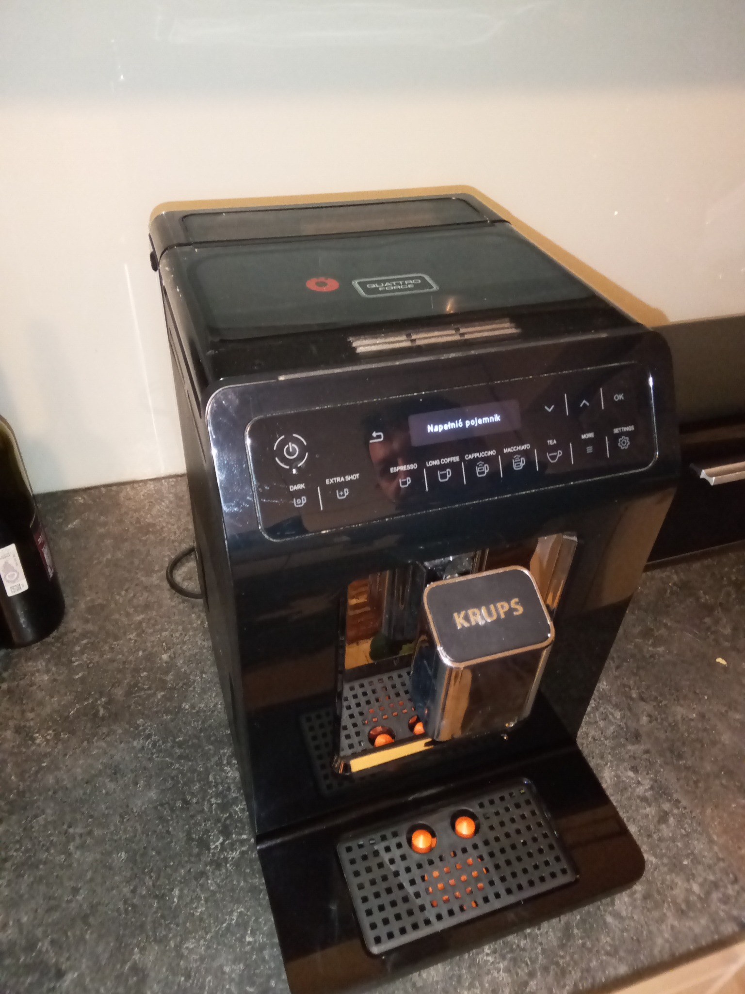 Czarny ekspres do kawy Krups na blacie, z komunikatem 'Napełnić pojemnik' na wyświetlaczu. Widoczny fragment butelki z boku. Oświetlenie górne.