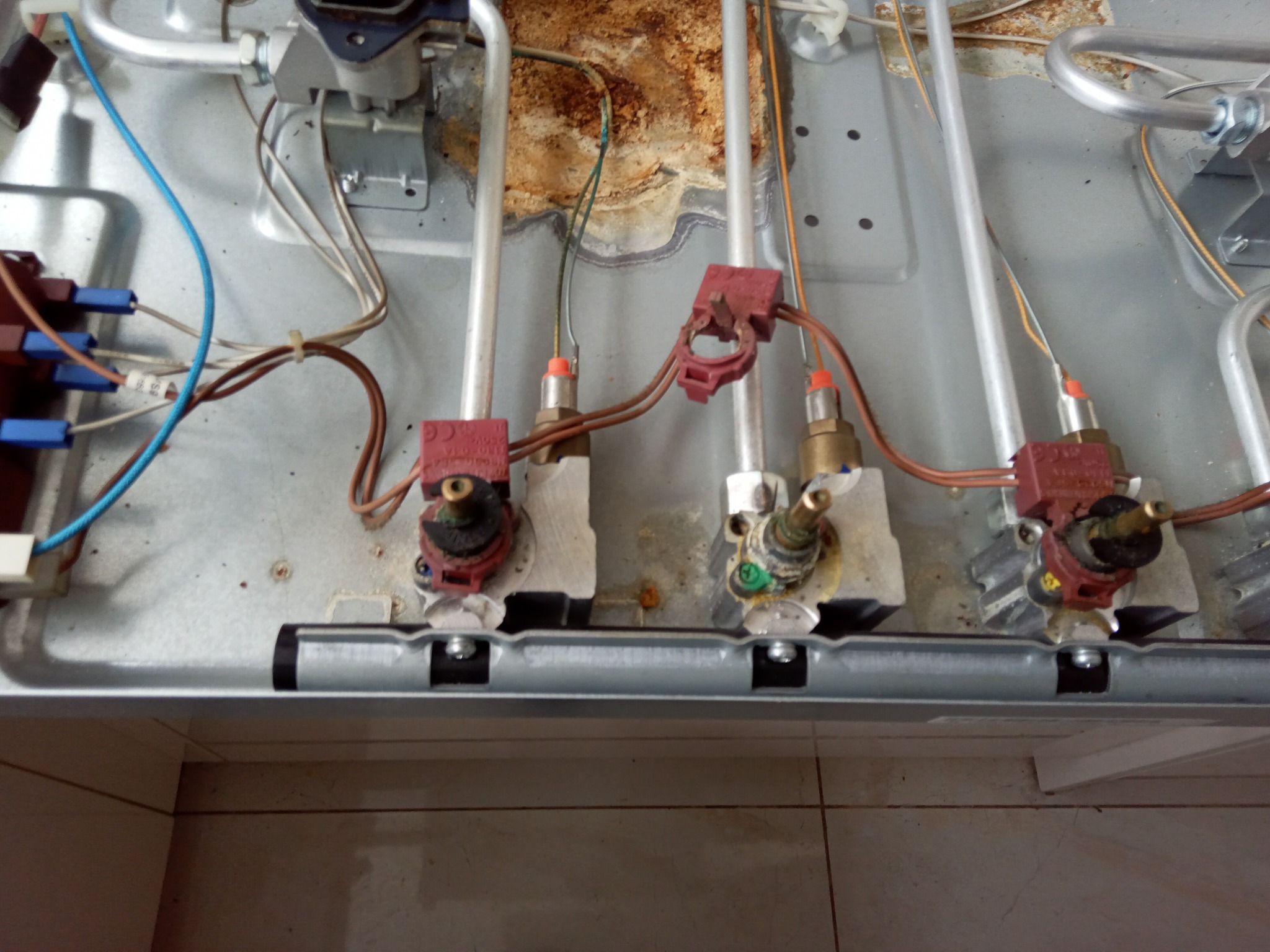 Wnętrze kuchenki gazowej z widocznymi elementami instalacji gazowej i elektrycznej, w trakcie naprawy lub serwisu. Widoczne ślady użytkowania i zabrudzenia.