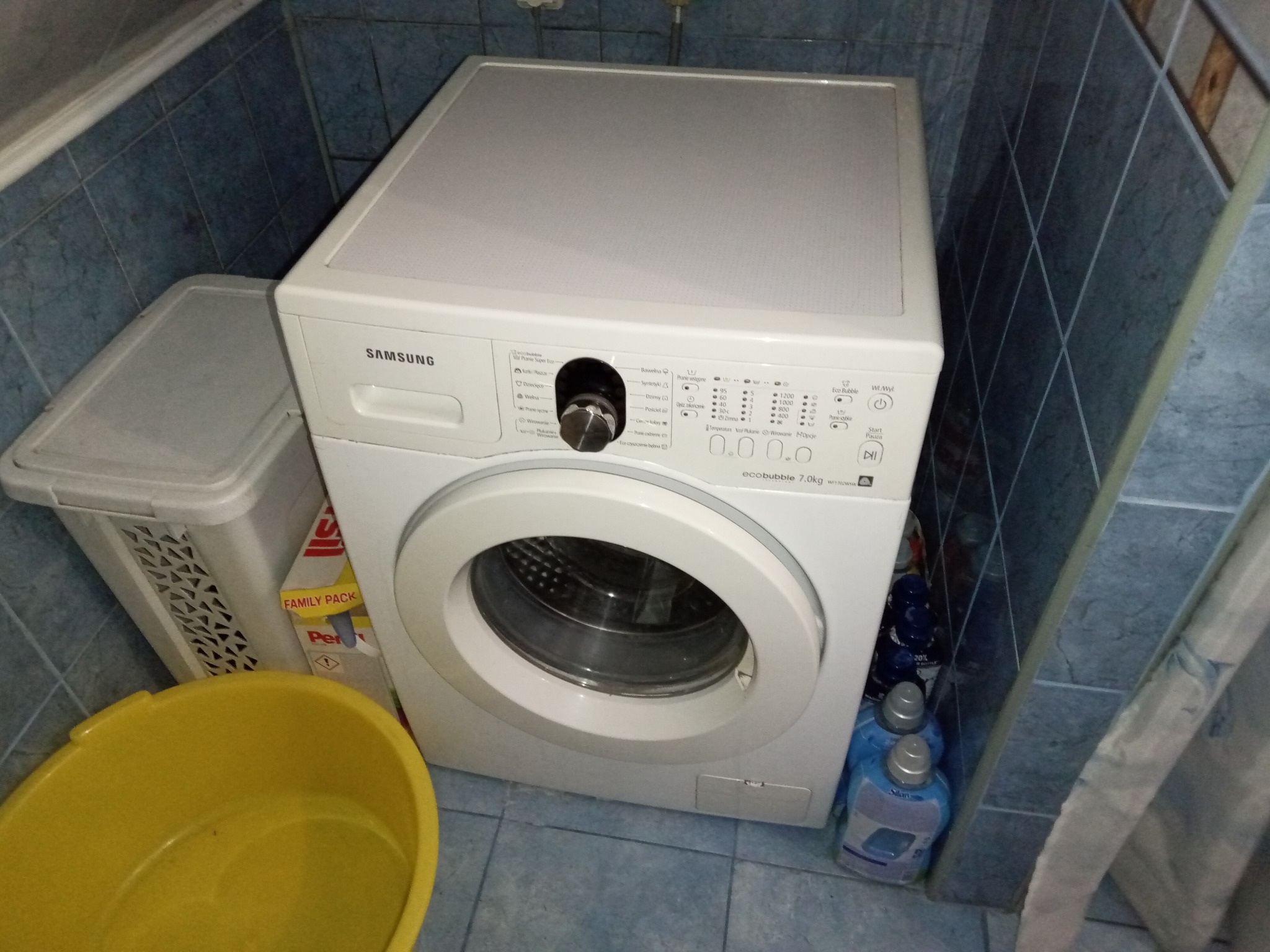 Biała pralka Samsung 7.0kg w łazience z niebieskimi płytkami, obok kosz na pranie i żółta miska. Widoczne środki piorące. Ujęcie z góry.