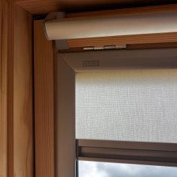 FAT CAP MARCIN ZEMLAK - Zamknięta roleta okienna marki Velux w drewnianej ramie, widoczna faktura materiału rolety i mechanizm zwijania. Stonowana kolorystyka, zbliżenie na detal.