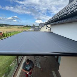 FAT CAP MARCIN ZEMLAK - Montaż szarej rolety tarasowej z czarną ramą na tle błękitnego nieba. Widoczny mężczyzna przy drabinie i krajobraz za domem.