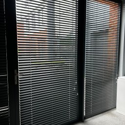 FAT CAP MARCIN ZEMLAK - Ciemne żaluzje aluminiowe zamontowane w oknie, z widokiem na ceglany budynek na zewnątrz. Delikatne światło słoneczne podkreśla strukturę żaluzji.