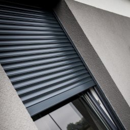 FAT CAP MARCIN ZEMLAK - Grafitowa roleta zewnętrzna częściowo opuszczona na oknie budynku o szarej, chropowatej elewacji. Widoczny fragment szyby i ramy okiennej.