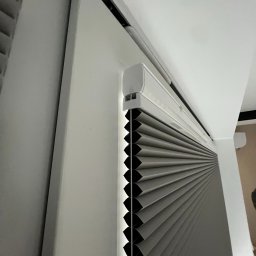 FAT CAP MARCIN ZEMLAK - Szare plisy okienne zamontowane w białej ramie okiennej, widoczne z bliska pod kątem. Charakterystyczna faktura plis i minimalistyczny design.