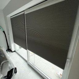 FAT CAP MARCIN ZEMLAK - Zasłonięte okno plisami w Gdańsku. Dwie szare plisy zamontowane w oknie balkonowym. Widoczne poziome paski materiału i białe ramy okienne.