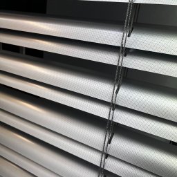 FAT CAP MARCIN ZEMLAK - Szczegółowe ujęcie aluminiowych żaluzji poziomych w kolorze srebrnym, z widocznym mechanizmem sterującym. Perforowana powierzchnia lameli.