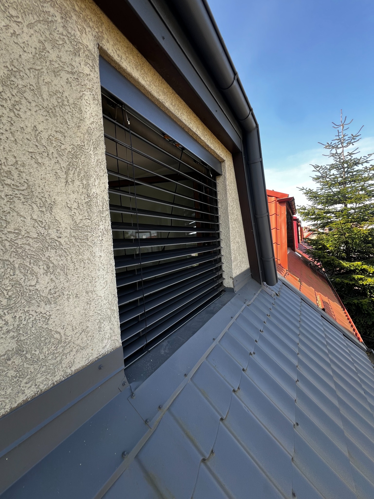 Zamknięte, czarne żaluzje aluminiowe zamontowane w oknie na poddaszu domu z szarą blachodachówką i beżową elewacją w słoneczny dzień. Widok z dołu.