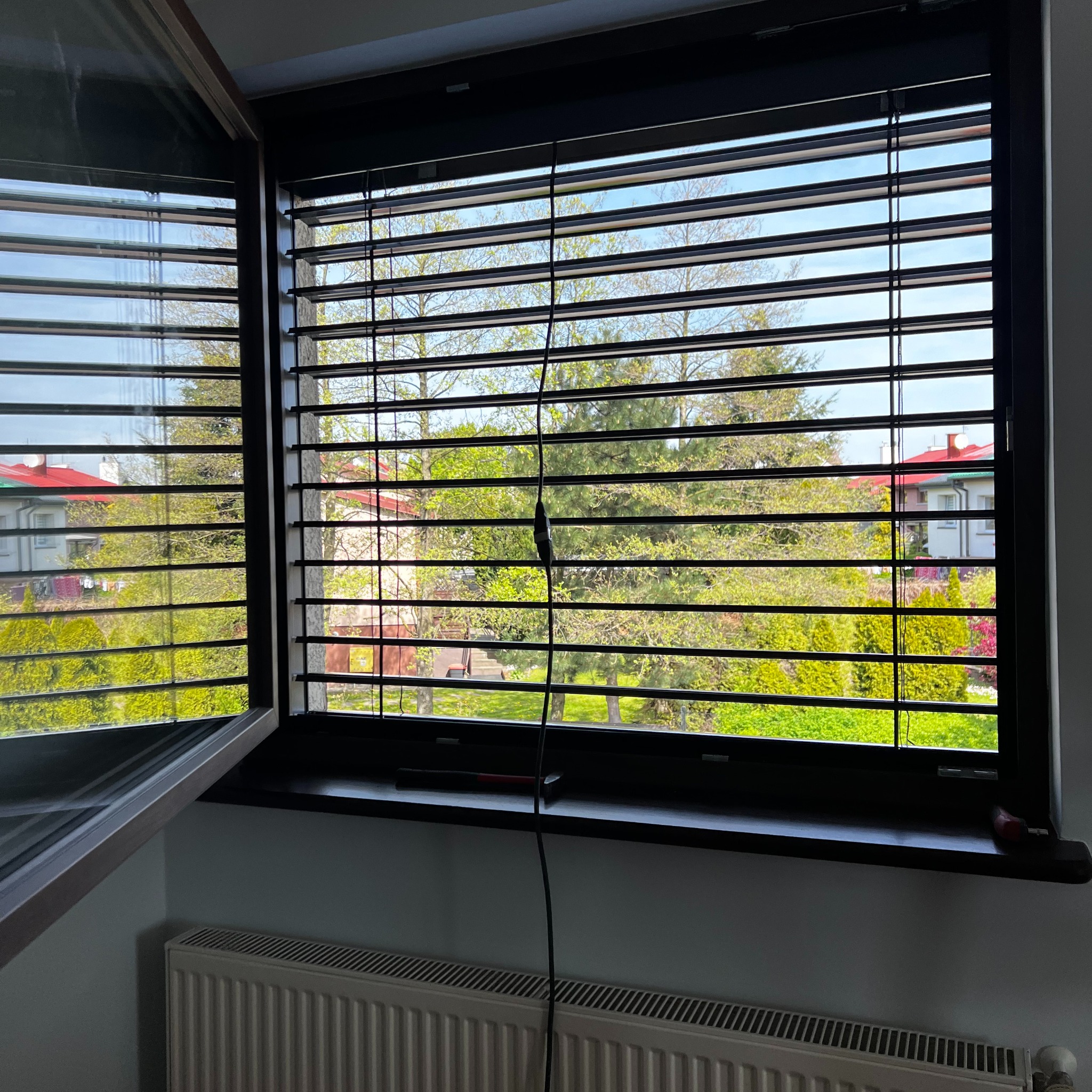 Ciemne żaluzje poziome w oknie z widokiem na zieleń. Jedno skrzydło okna otwarte, na parapecie leży młotek. Widoczny grzejnik pod oknem.