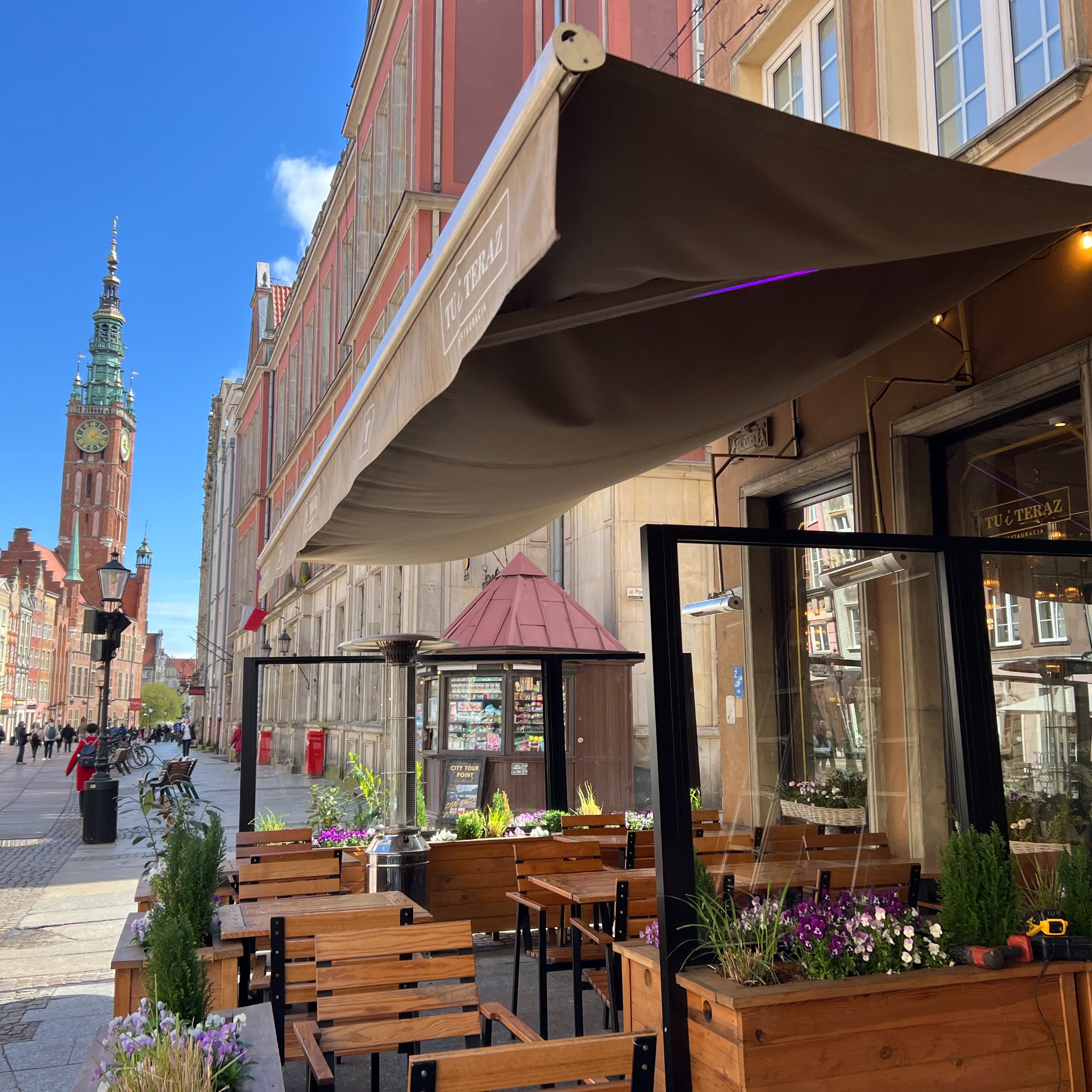 Zewnętrzny ogródek restauracji z rozłożoną markizą w słoneczny dzień. W tle zabytkowa architektura Gdańska i wieża Ratusza Głównego Miasta.