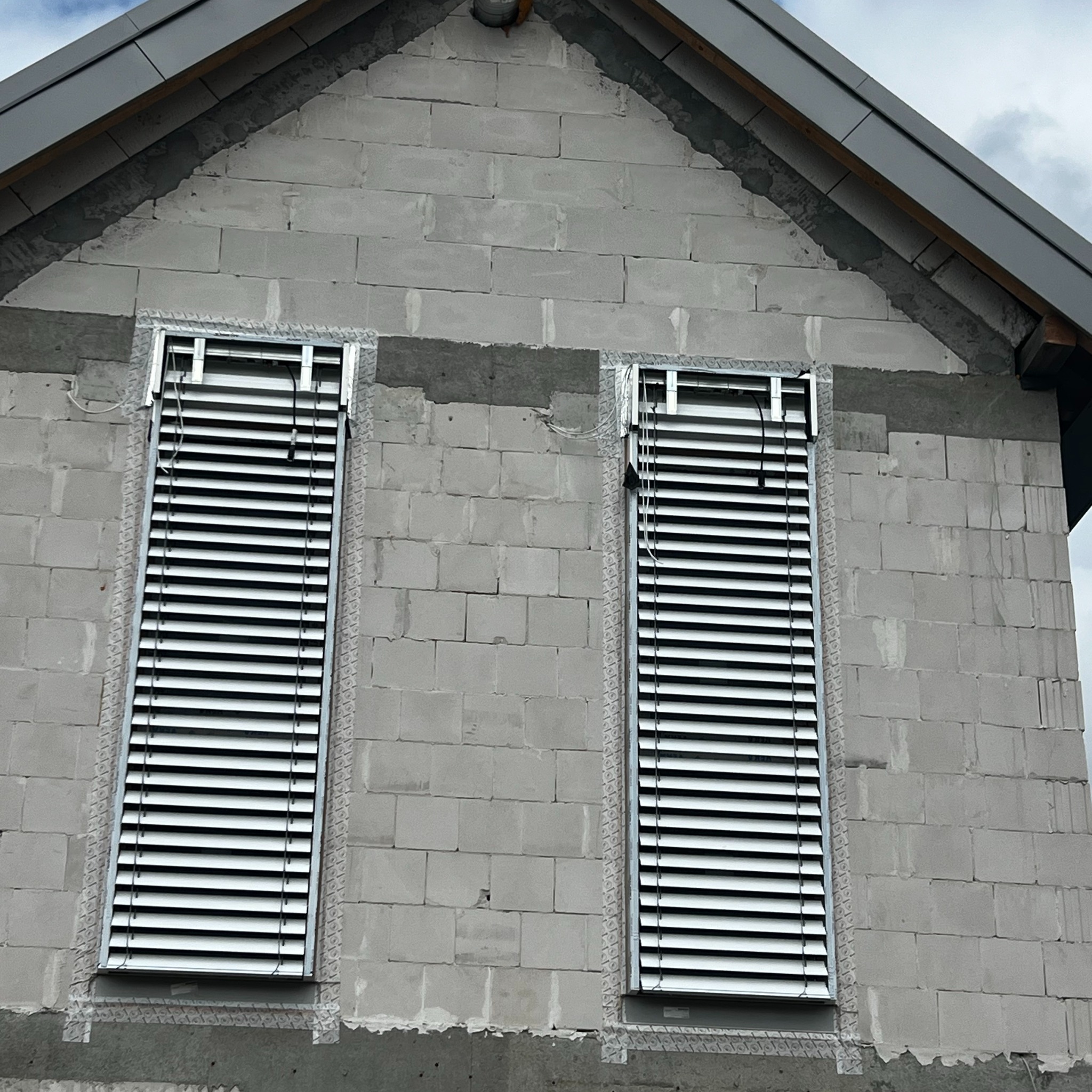 Dwa okna z zamontowanymi, aluminiowymi żaluzjami zewnętrznymi na surowej ścianie budynku z betonu komórkowego. Widoczne elementy montażowe.