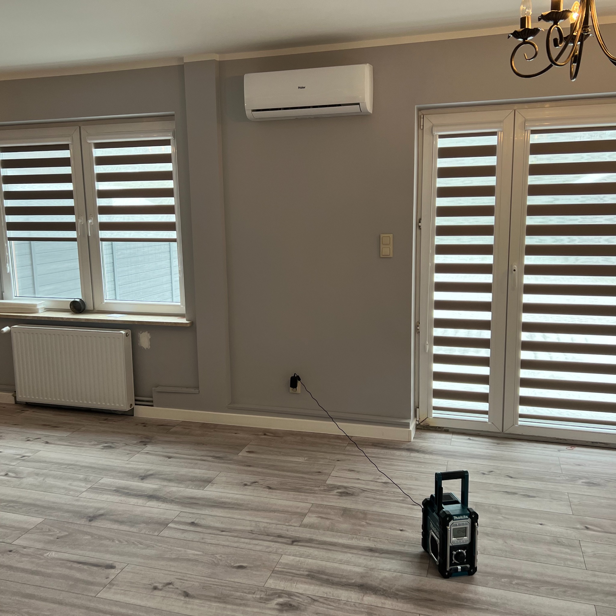 Wnętrze z roletami dzień noc w oknach i drzwiach balkonowych, klimatyzacja na ścianie, podłoga drewniana, radio budowlane na podłodze.