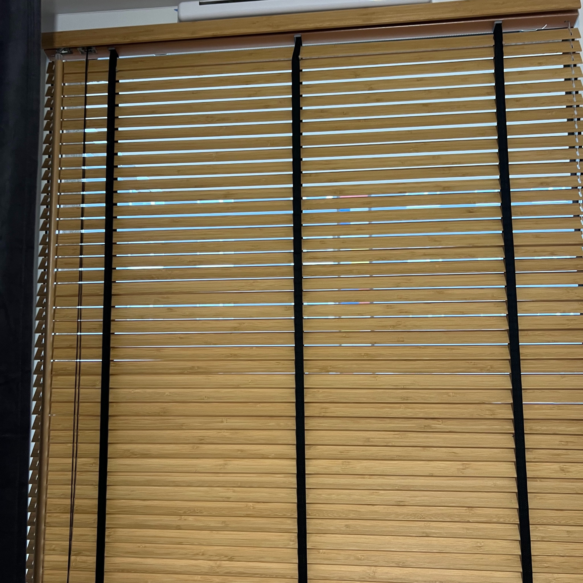 Zamknięte żaluzje drewniane w odcieniach beżu z czarnymi taśmami, widoczne poziome lamele i fragment okna w tle. Delikatne światło dzienne.