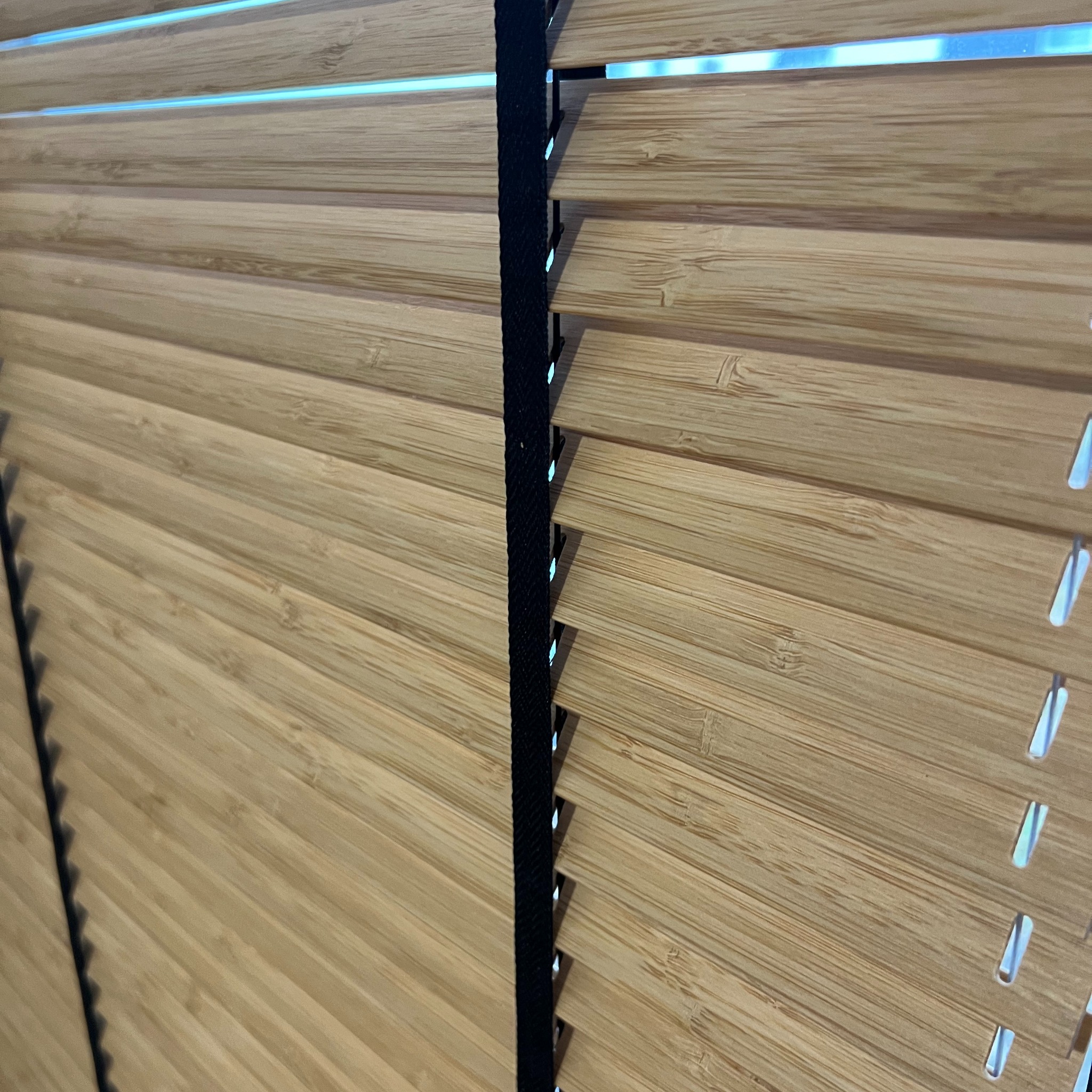 Drewniane żaluzje poziome z czarnym sznurkiem, delikatnie uchylone, wpuszczające subtelne światło. Widoczna faktura drewna i precyzyjne wykonanie.