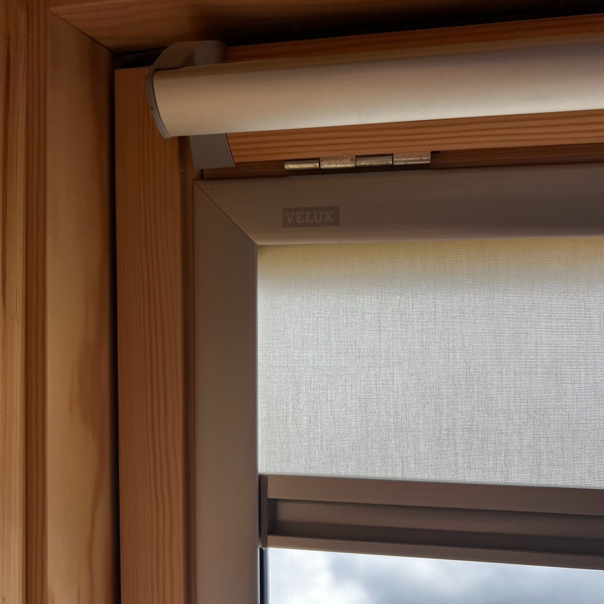 Zamknięta roleta okienna marki Velux w drewnianej ramie, widoczna faktura materiału rolety i mechanizm zwijania. Stonowana kolorystyka, zbliżenie na detal.