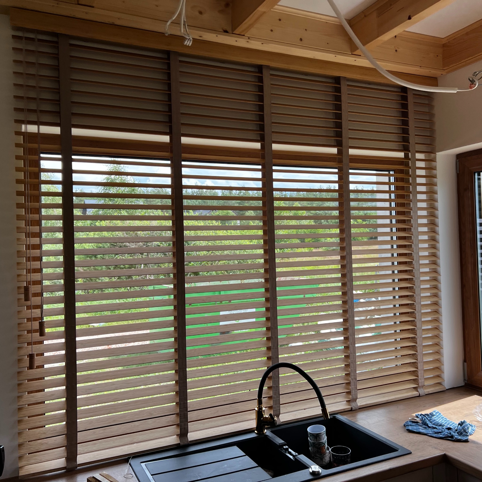 Jasne żaluzje drewniane zamontowane w oknie nad zlewem kuchennym. Widok na zieleń za oknem. Elementy drewniane w kolorze naturalnego drewna.