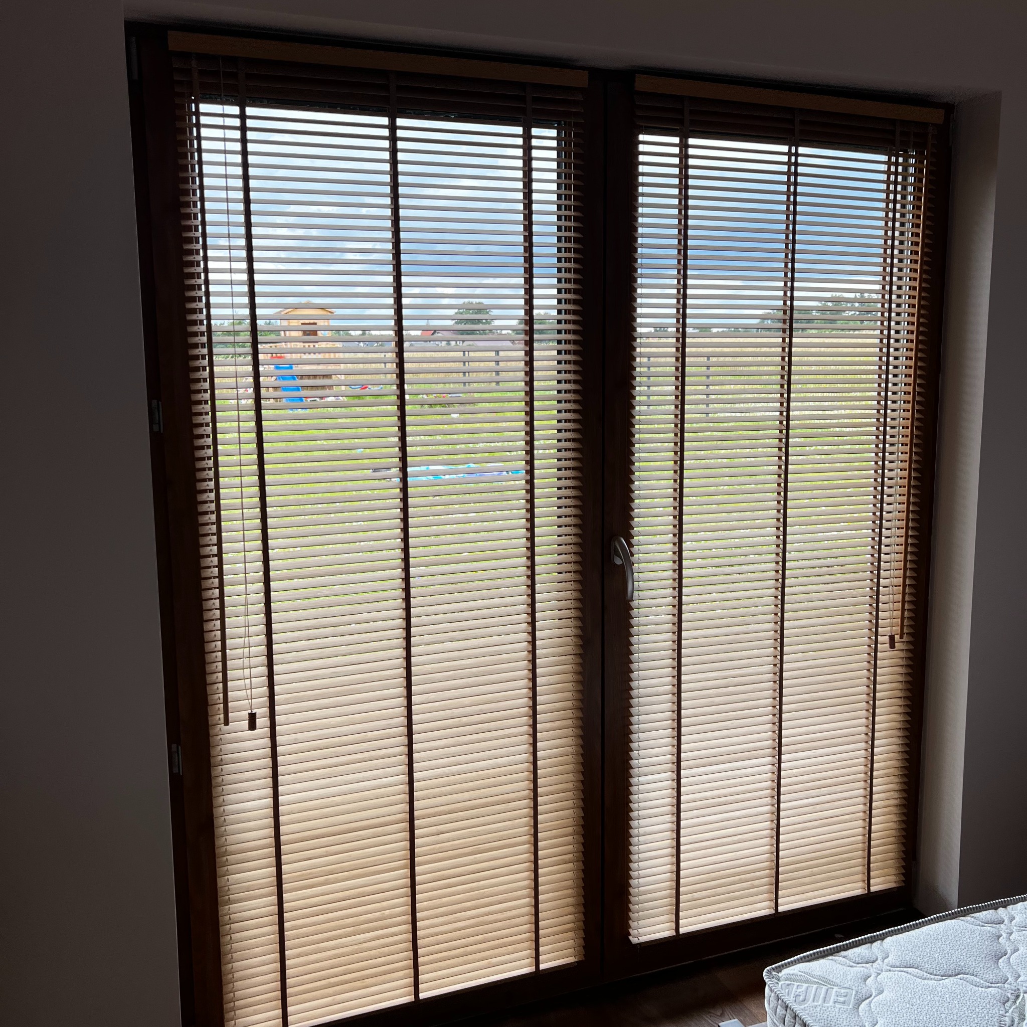 Drewniane żaluzje poziome w oknie z widokiem na plac zabaw. Ciepłe światło podkreśla strukturę drewna. Widoczne sznurki regulacyjne i fragment materaca.