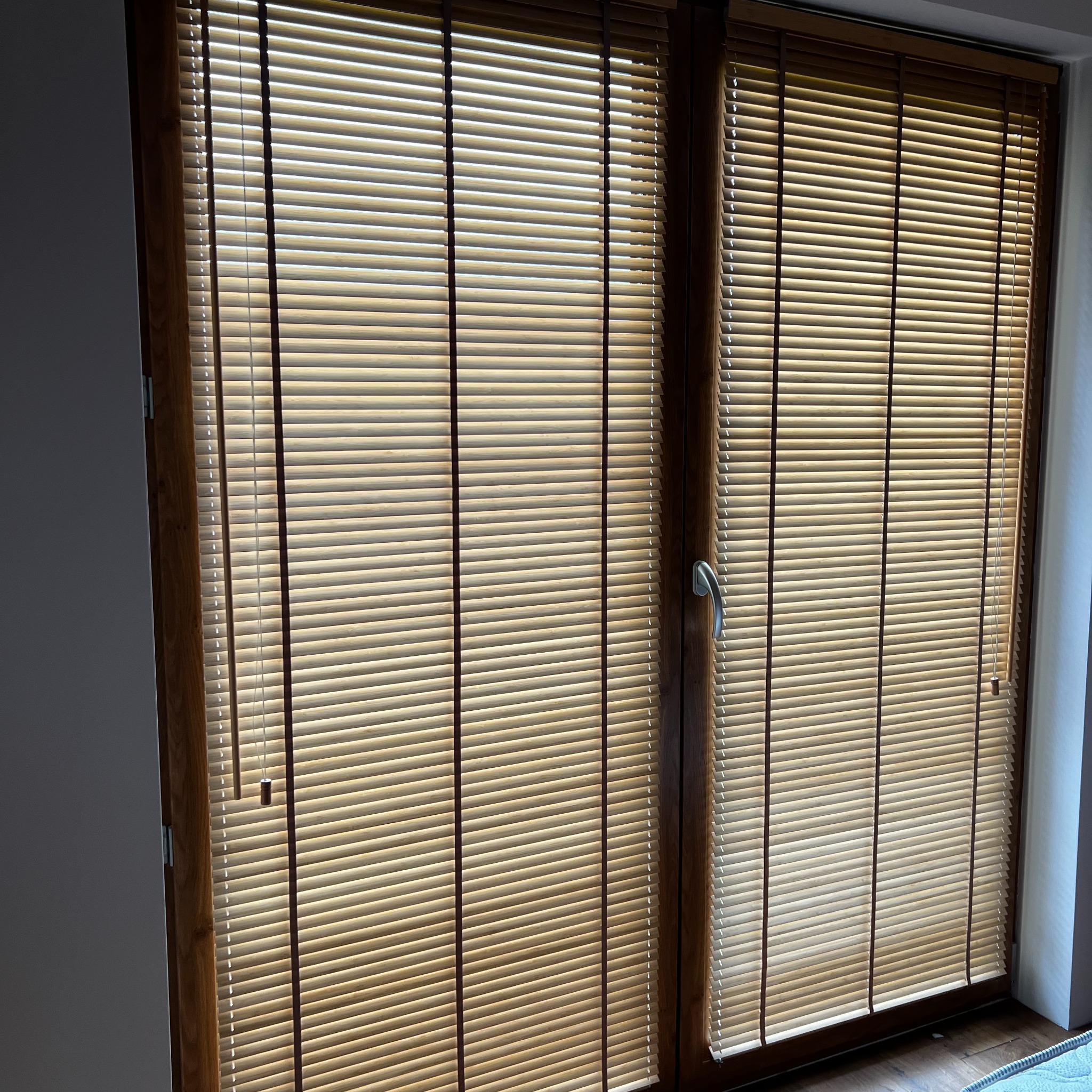 Drewniane żaluzje poziome w oknie z brązową ramą, delikatnie przepuszczające światło słoneczne. Widoczne sznurki do regulacji po lewej stronie.