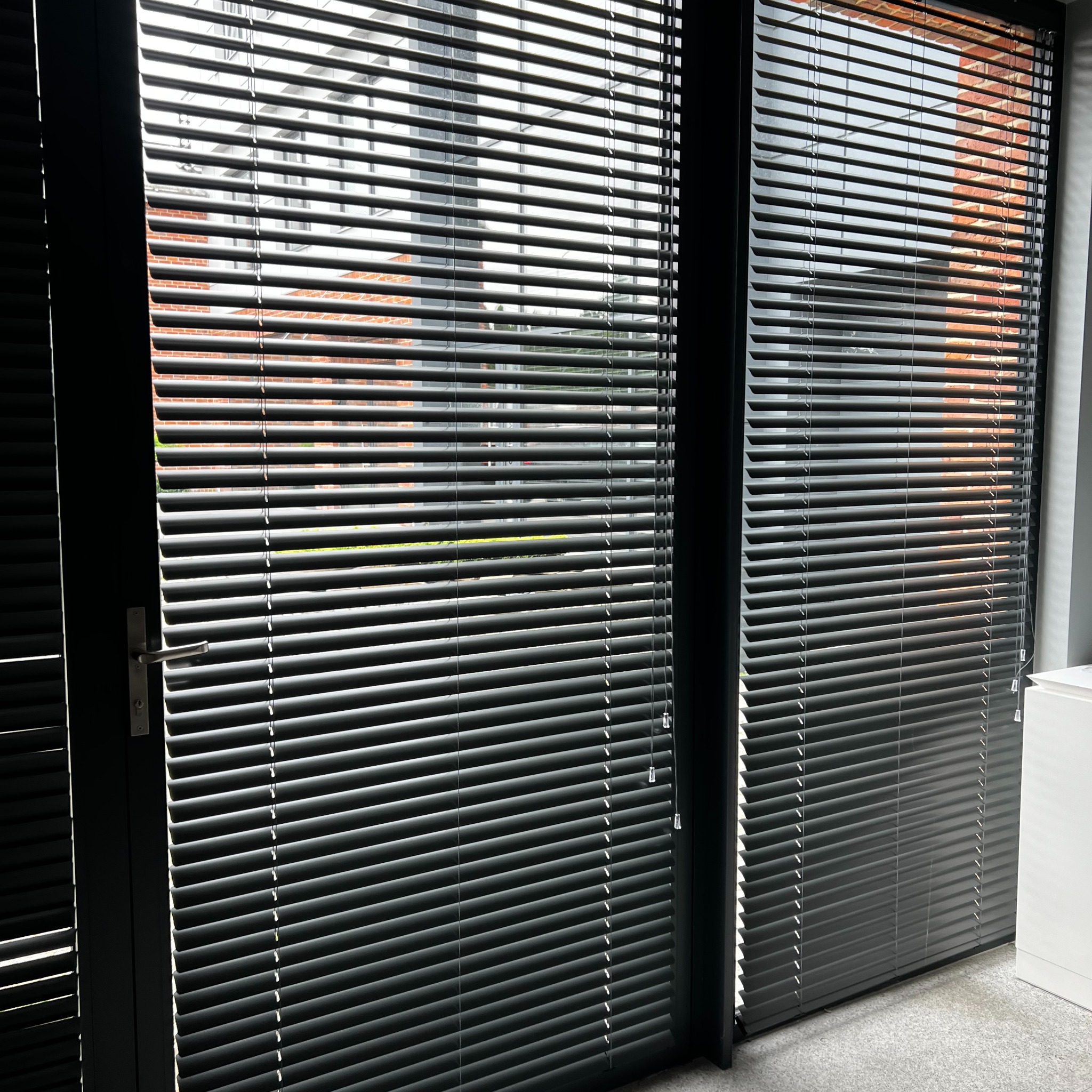 Ciemne żaluzje aluminiowe zamontowane w oknie, z widokiem na ceglany budynek na zewnątrz. Delikatne światło słoneczne podkreśla strukturę żaluzji.
