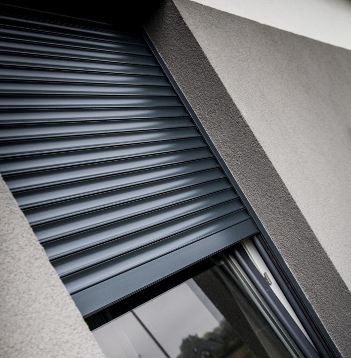 Grafitowa roleta zewnętrzna częściowo opuszczona na oknie budynku o szarej, chropowatej elewacji. Widoczny fragment szyby i ramy okiennej.