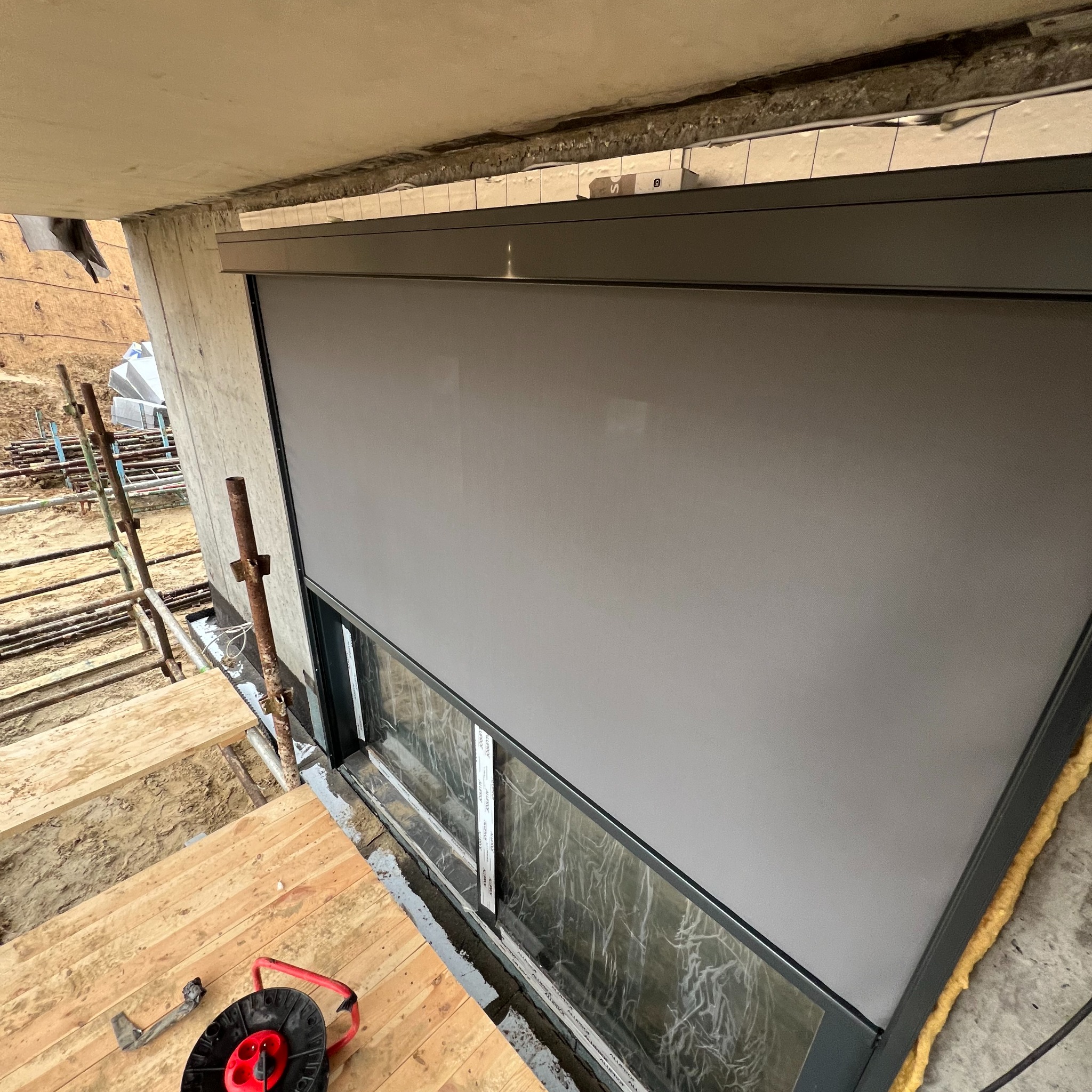 Szara roleta zewnętrzna zamontowana w oknie budynku w trakcie budowy, widok z góry na drewniany podest rusztowania i zwinięty przedłużacz.