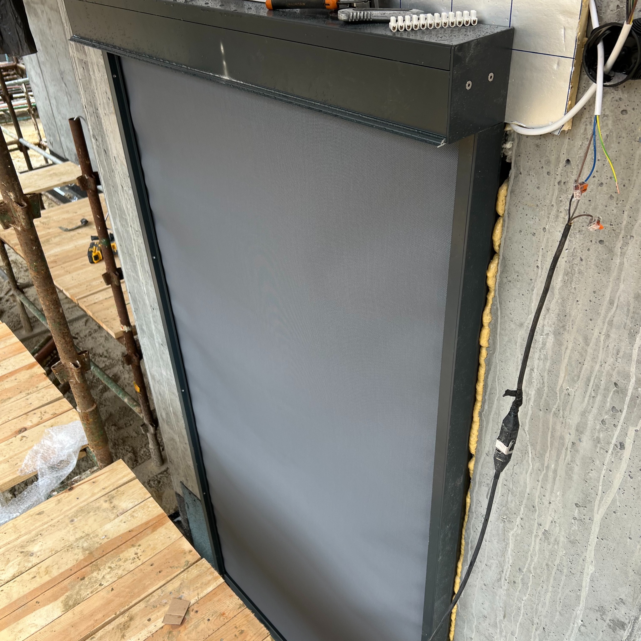 Szara roleta zewnętrzna zamontowana na elewacji budynku w trakcie prac wykończeniowych, widoczne rusztowania i narzędzia.