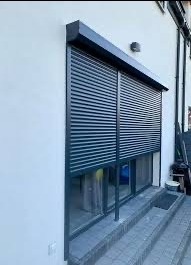 Grafitowa roleta zewnętrzna, zamontowana na oknie tarasowym w nowoczesnym domu. Widoczne schody prowadzące do okna i fragment elewacji.