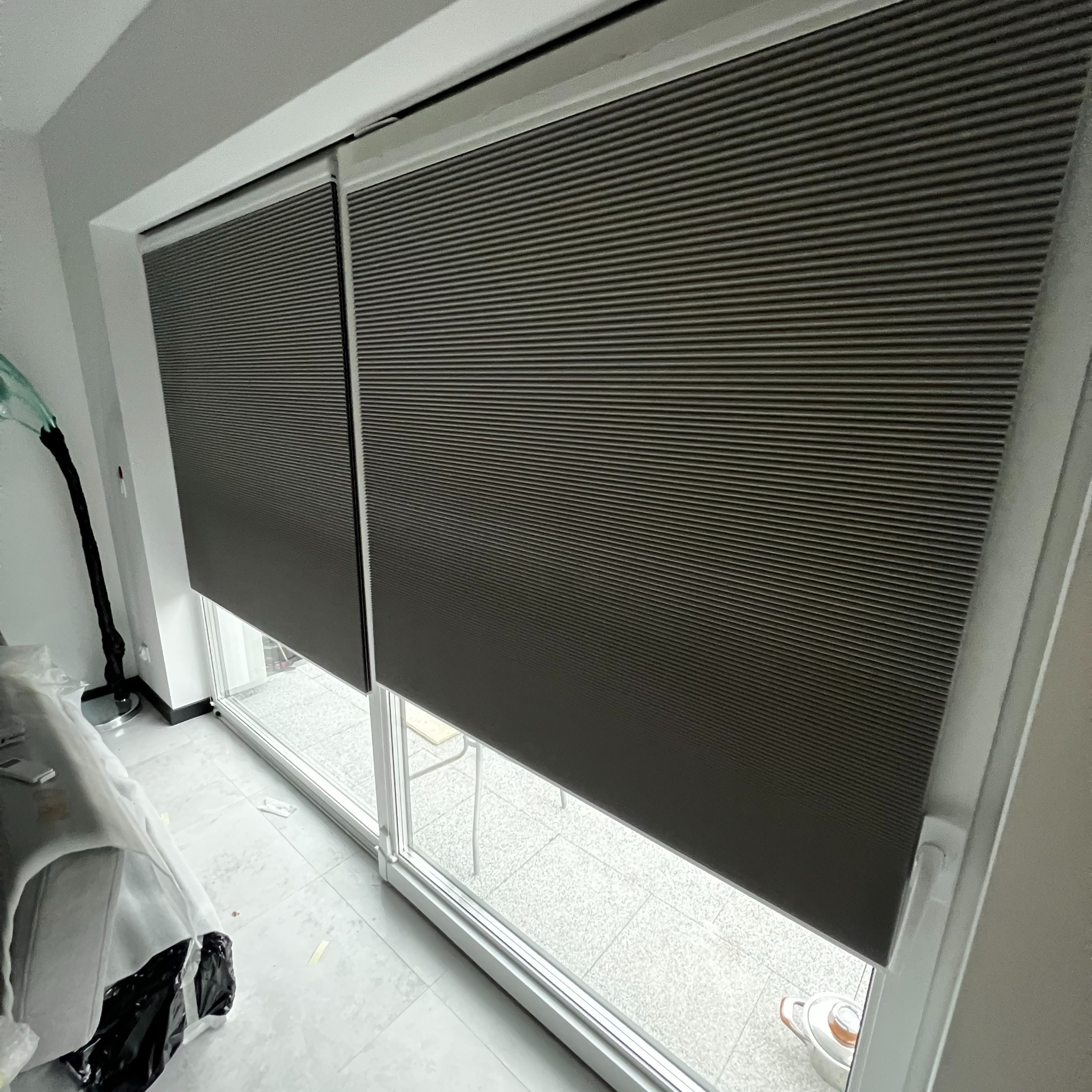 Zasłonięte okno plisami w Gdańsku. Dwie szare plisy zamontowane w oknie balkonowym. Widoczne poziome paski materiału i białe ramy okienne.