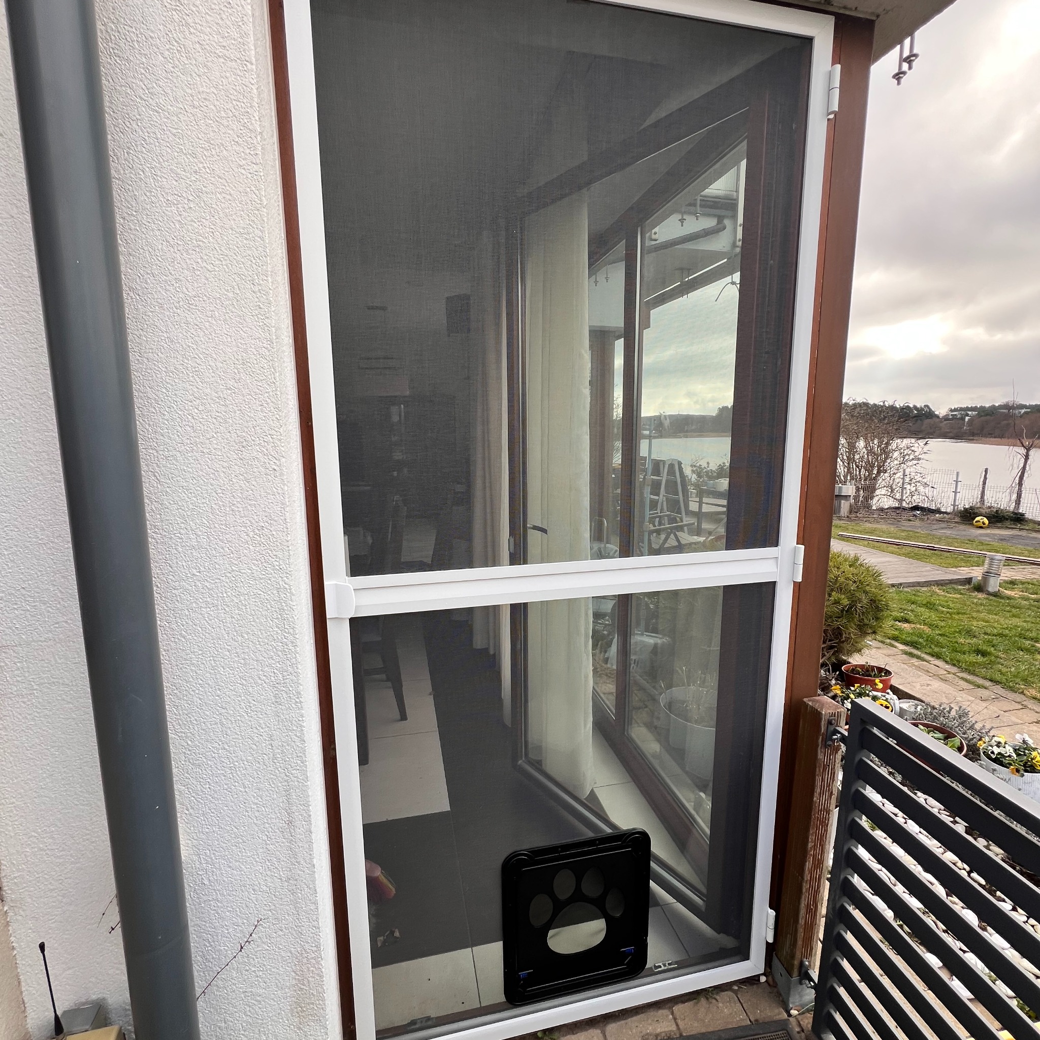 Biała moskitiera z klapką dla zwierząt na drzwiach balkonowych, widok z zewnątrz. W tle zieleń ogrodu i jezioro. Detal: czarna klapka z odciskiem łapy.